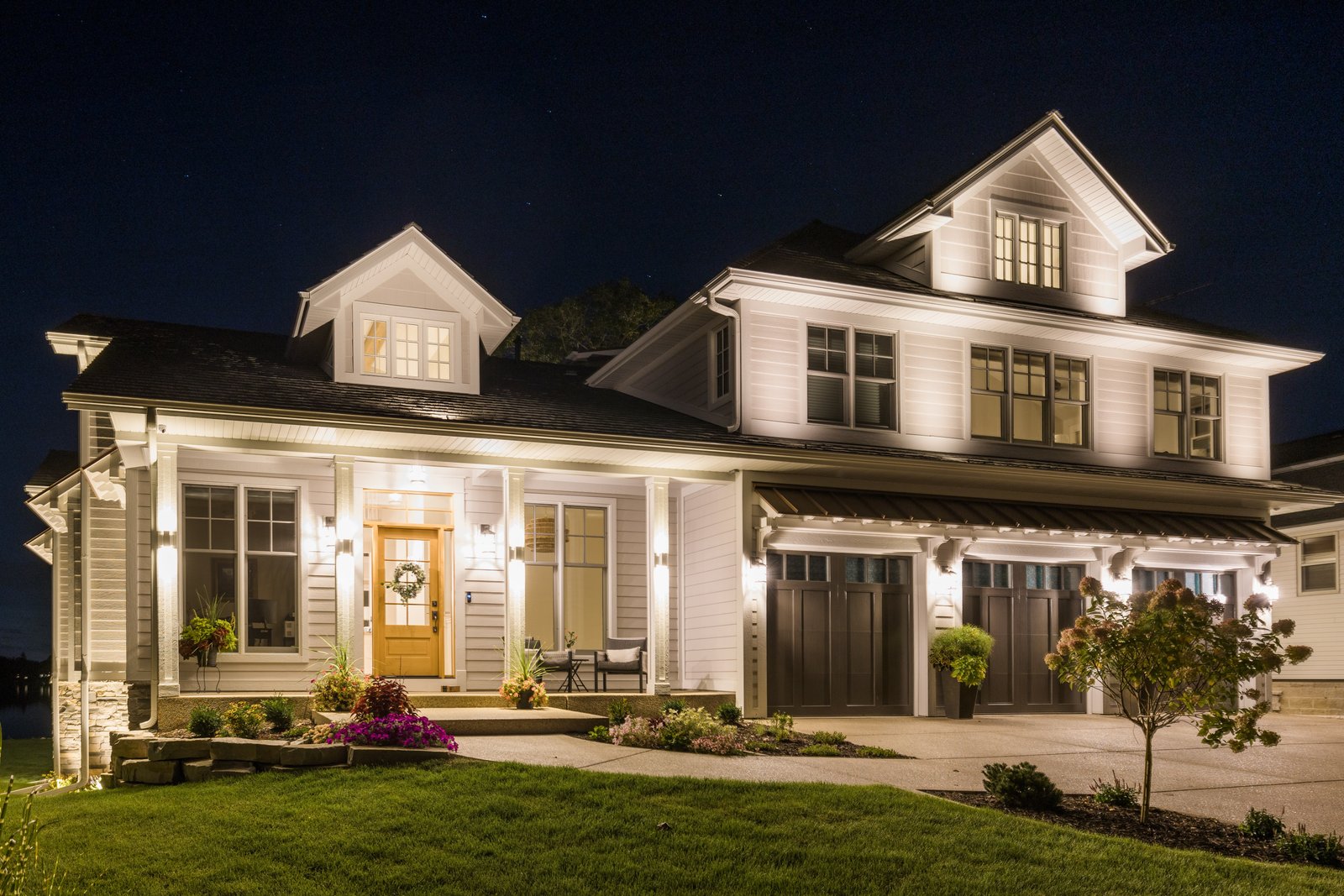 Front elevation at dusk under a starry sky: lit porch with a wood front door, dormers, and a three-car garage with carriage doors
