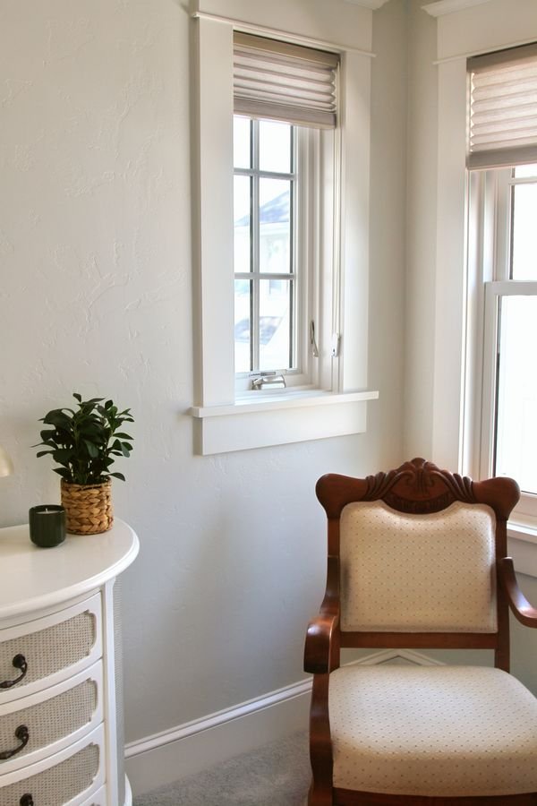 Bedroom corner with an antique armchair, a painted chest, and two windows with woven shades