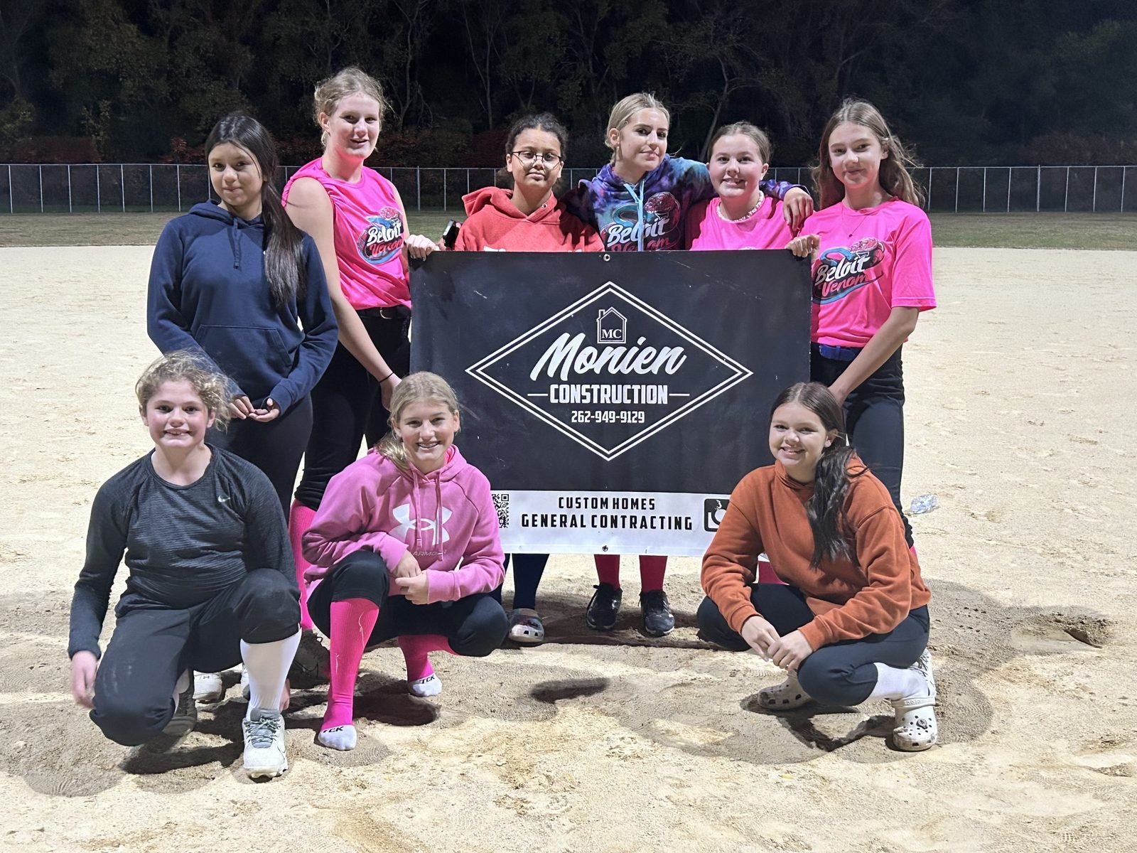 Players from the Beloit Venom softball team gathered on the infield at night, holding the Monien Construction banner
