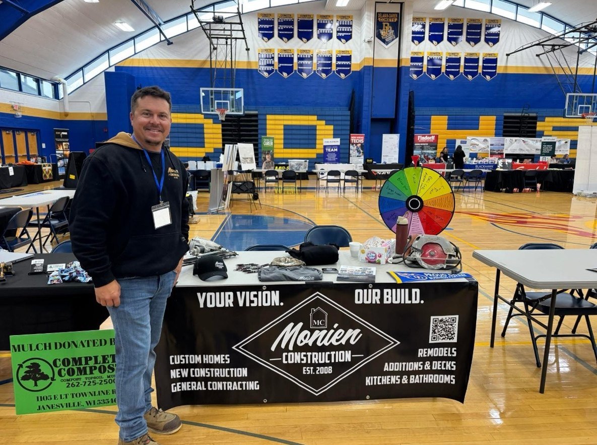 Nick Monien standing at the Monien Construction booth in the Delavan-Darien High School gym, with the company banner, tools, and a prize wheel on the table