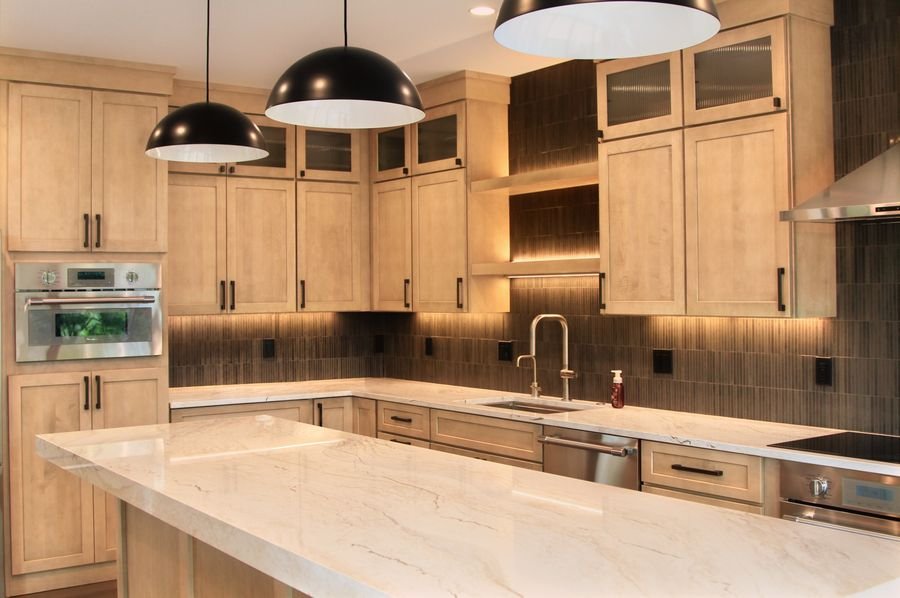 Long stone island under black dome pendants, maple cabinetry, and a dark tile backsplash lit from beneath the cabinets