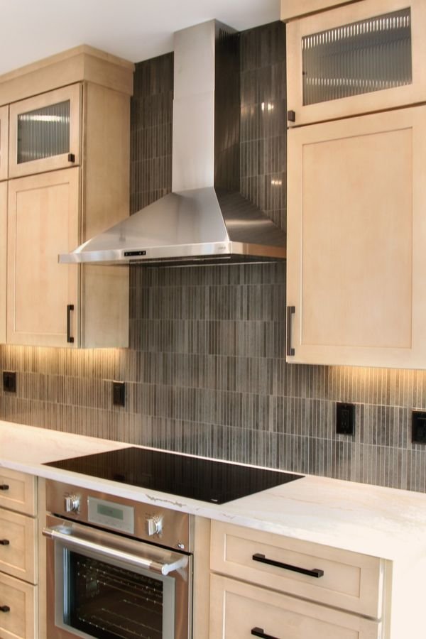 Stainless chimney hood over the cooktop, with dark vertical tile rising to the ceiling between maple cabinets
