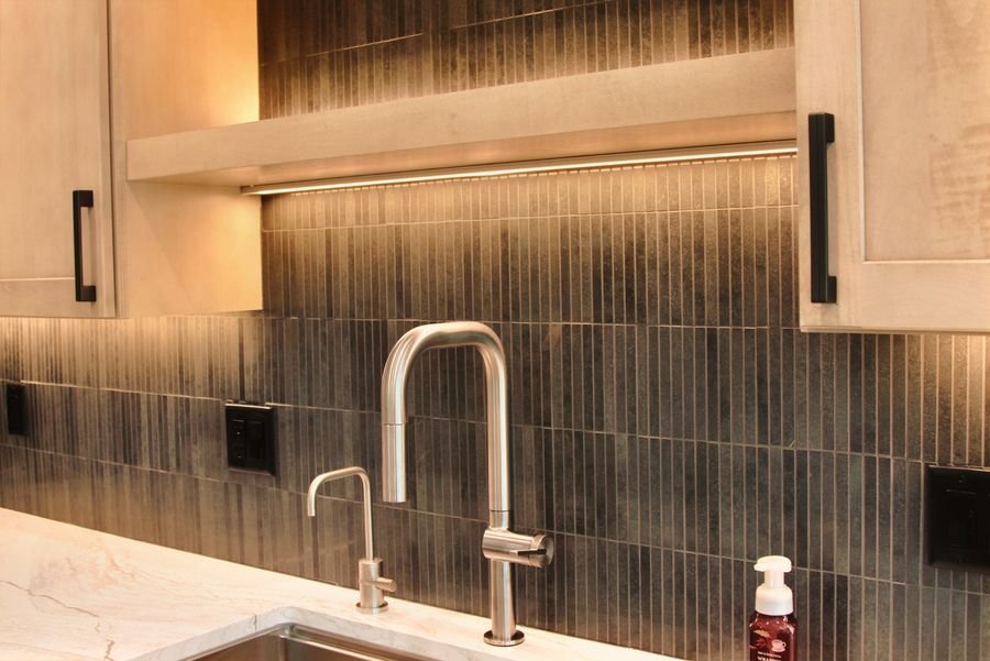 Pull-down faucet and filtered water tap in front of dark vertical tile lit by a strip beneath the floating shelf
