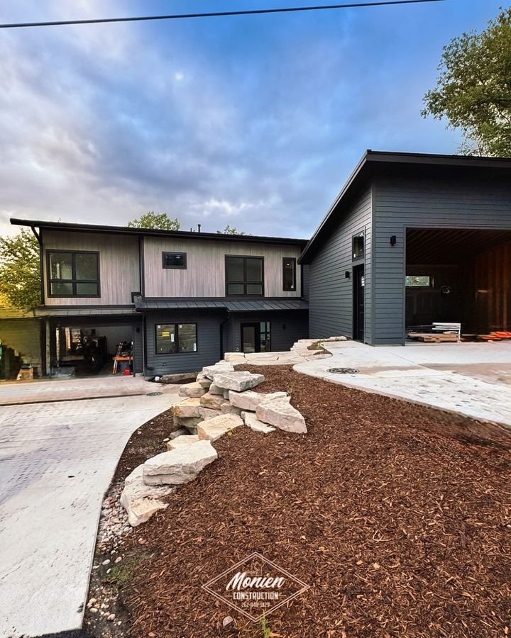 Front elevation with light wood siding above dark siding, the detached garage, and a natural stone outcrop beside the drive