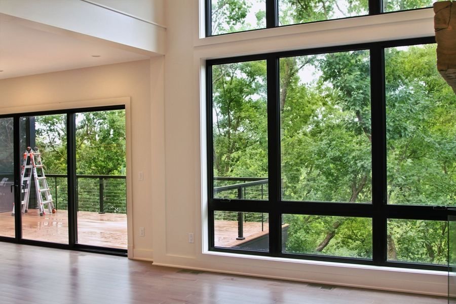 Two-story black-framed window wall and sliding door opening to the deck, with the trees and lake beyond